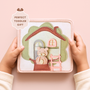 a fabric activity book for toddlers. The cover shows a house design with a tree-shaped background, a ladder, and a smiling teddy bear. The text "PERFECT TODDLER GIFT" is displayed on the top left.