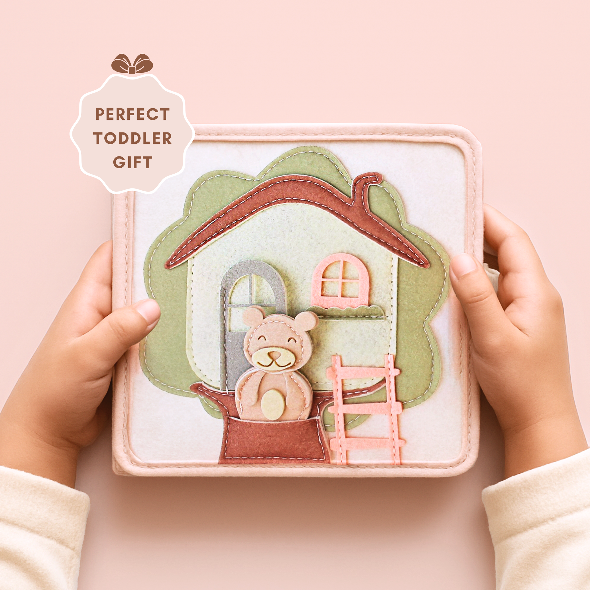 a fabric activity book for toddlers. The cover shows a house design with a tree-shaped background, a ladder, and a smiling teddy bear. The text "PERFECT TODDLER GIFT" is displayed on the top left.