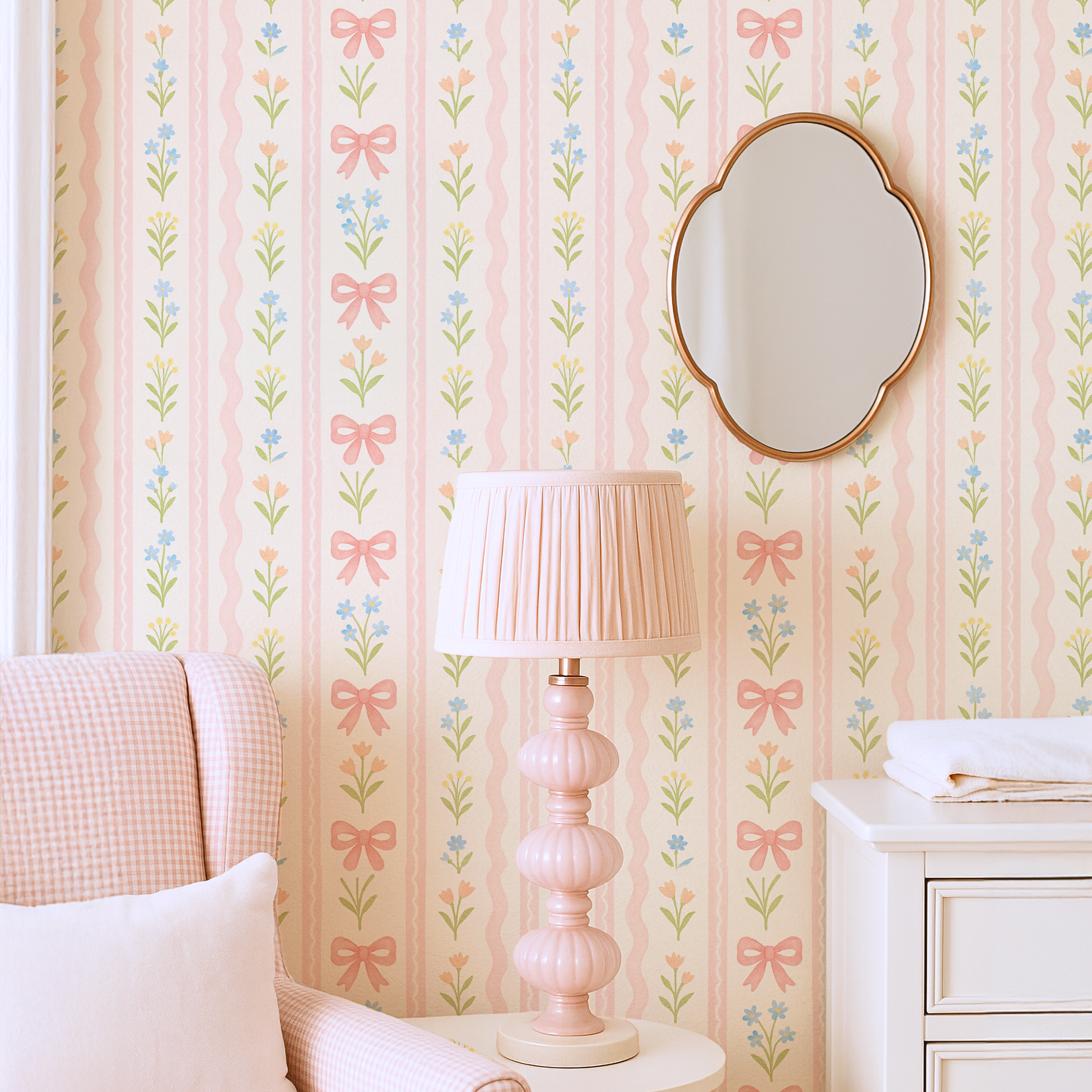 Blush nursery wall with pink bow stripe wallpaper, scalloped mirror and pleated table lamp styled in a soft feminine space.