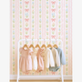 Pastel girls nursery wallpaper with bows and floral stripes behind baby clothes rack, soft pink aesthetic toddler bedroom decor